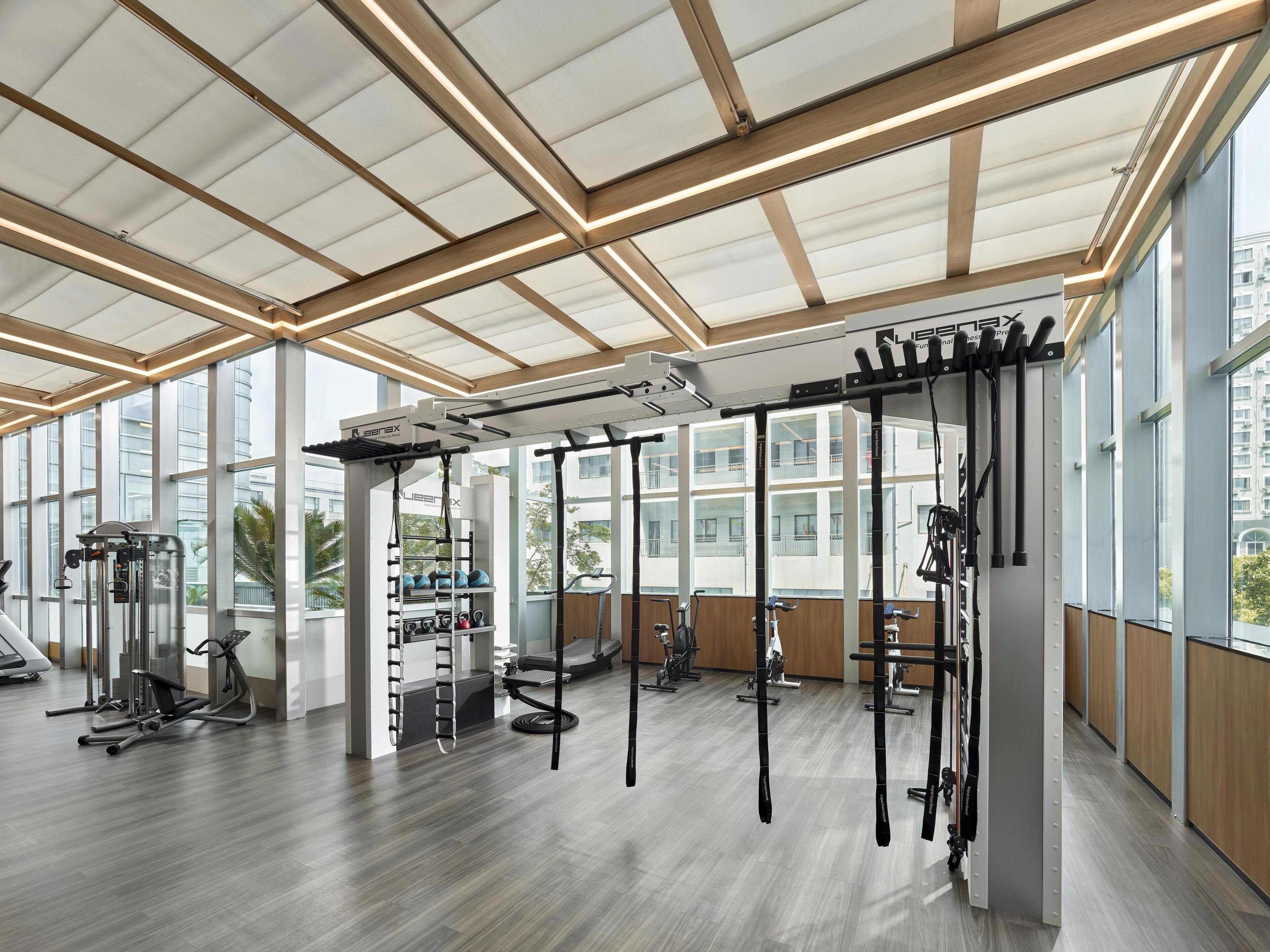 A modern gym with ceiling-mounted exercise equipment, large windows, and a wooden floor.