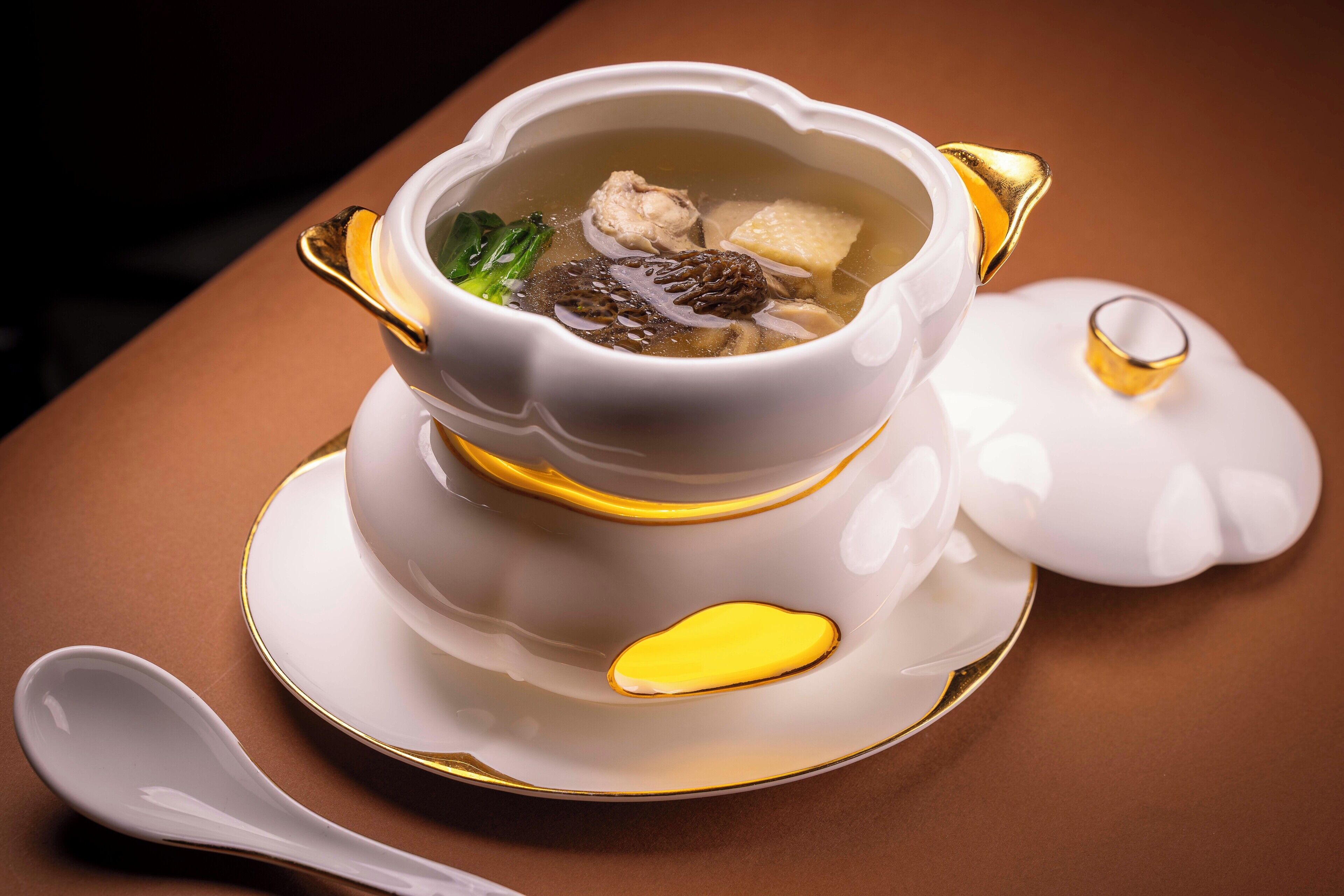 A white ceramic soup bowl with gold accents on a matching saucer, accompanied by a spoon and a small bowl.