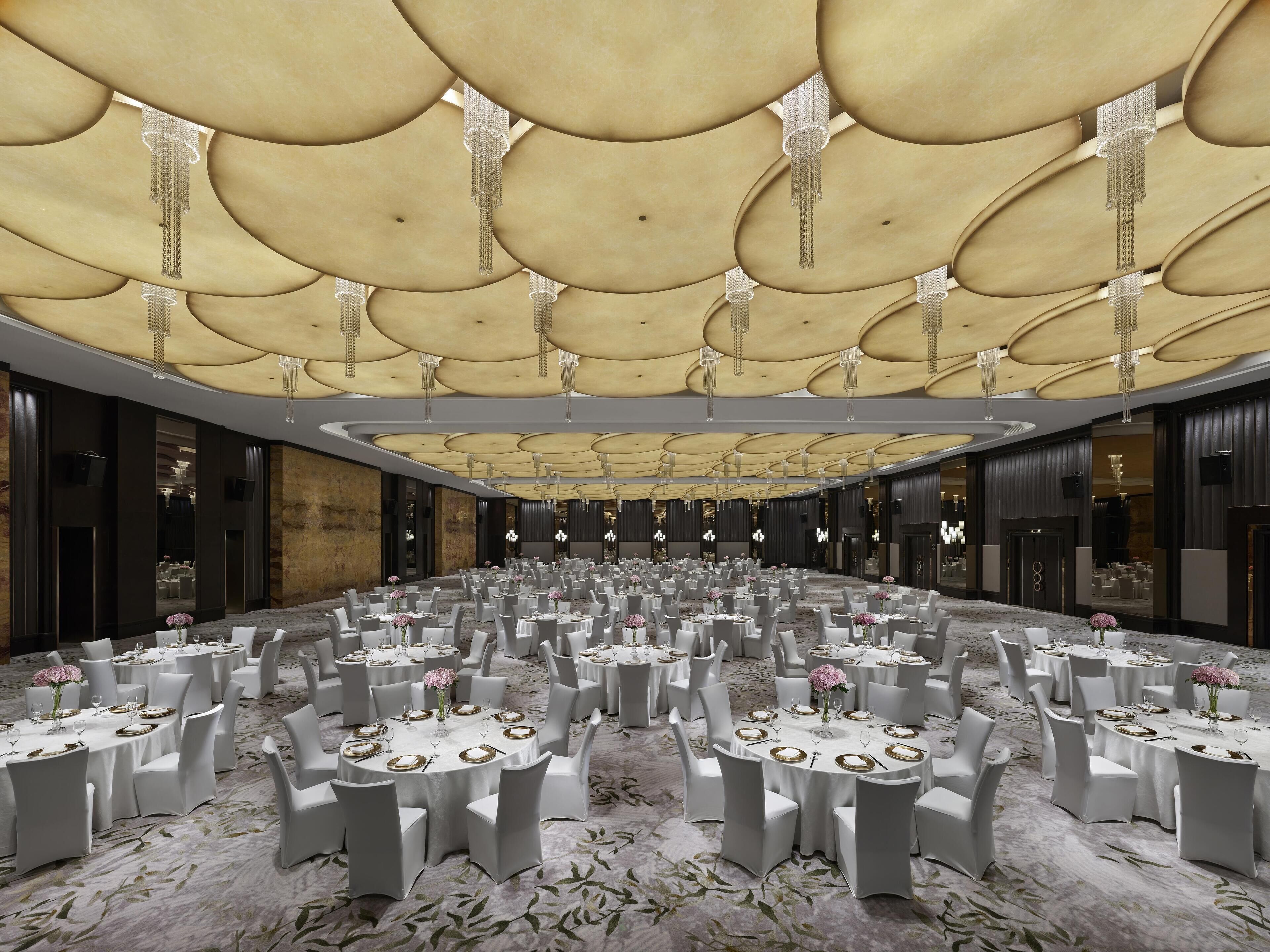 A spacious banquet hall with round tables set for dining, a high ceiling with decorative lighting, and a marble floor.