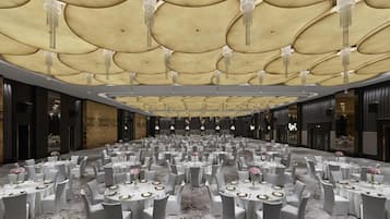 A spacious banquet hall with round tables set for dining, a high ceiling with decorative lighting, and a marble floor.