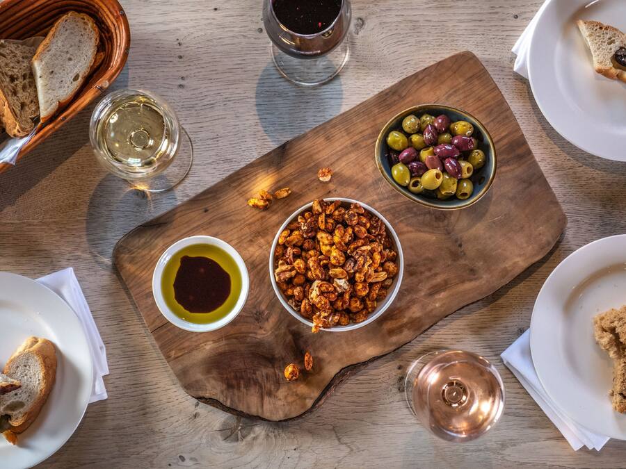 Una mesa de madera con diversos alimentos, como pan, aceitunas y frutos secos, junto con copas de vino.