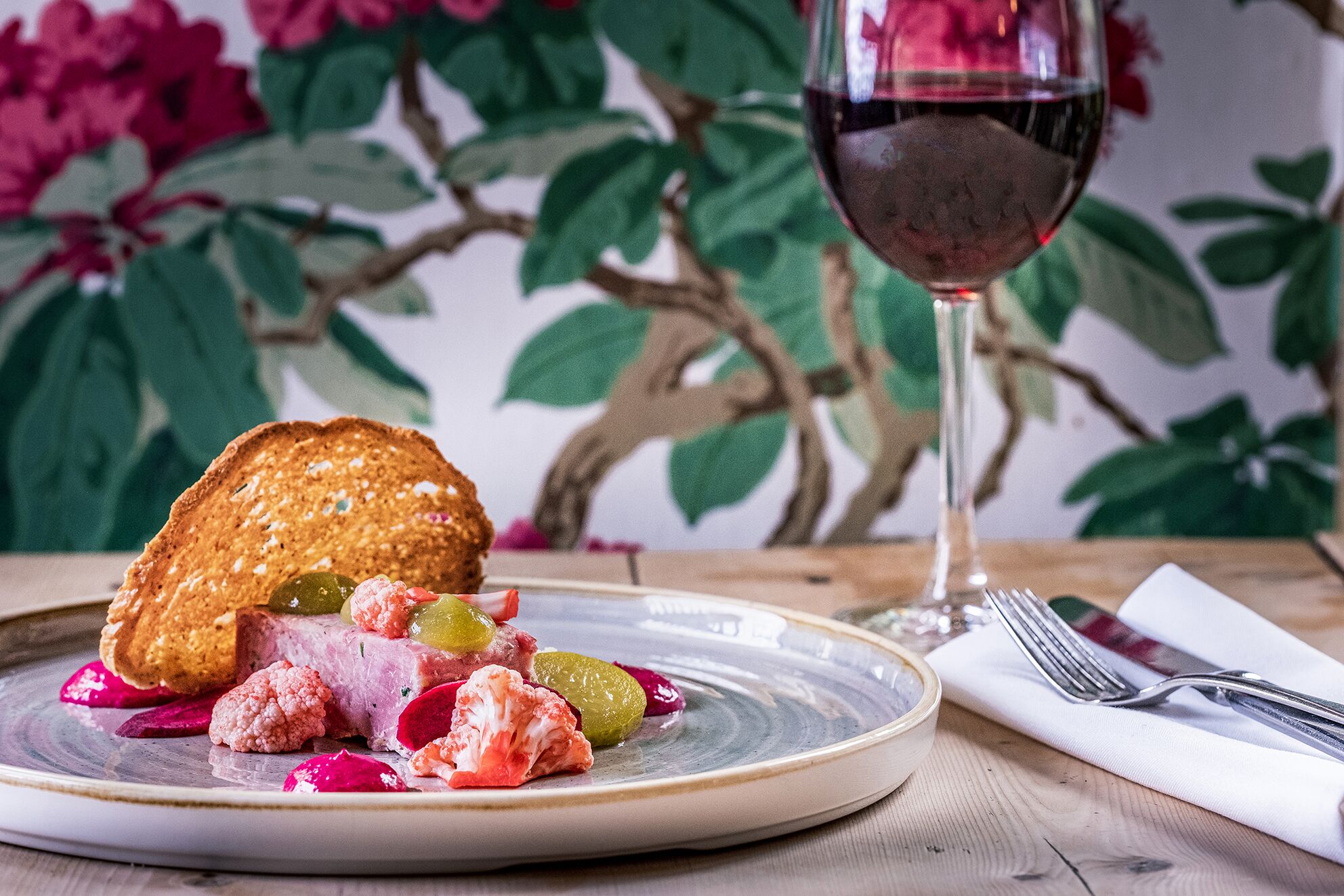 A plate with food and a glass of red wine on a table with a floral wallpaper background.