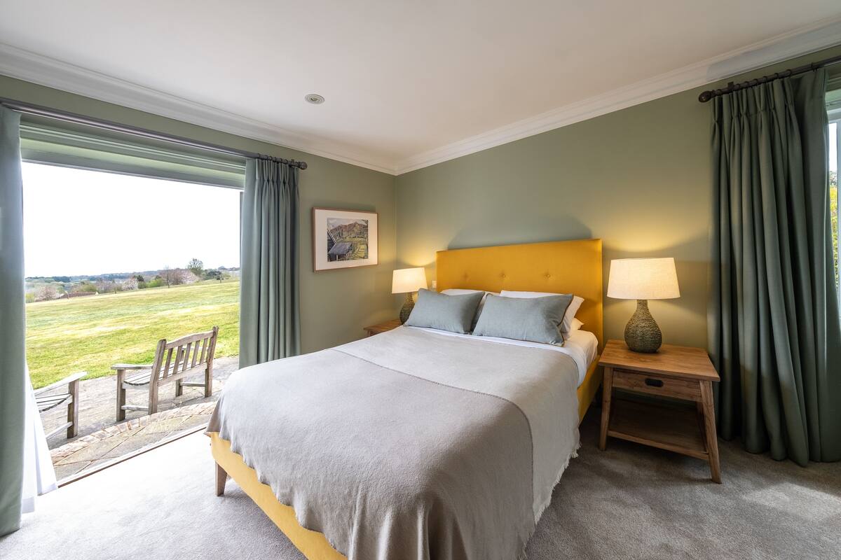 Een slaapkamer met een bed, twee nachtkastjes, een raam met gordijnen en uitzicht op een grasveld.