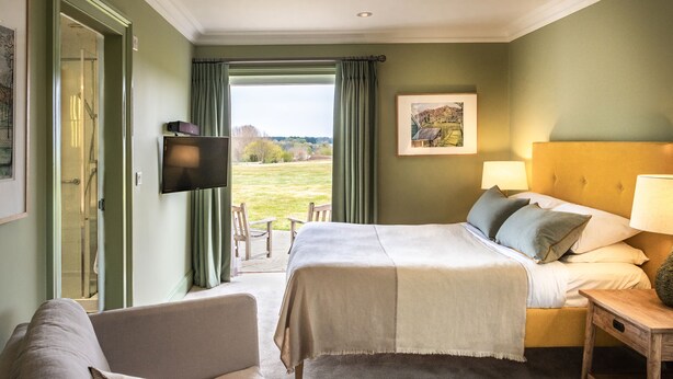 Een slaapkamer met een bed, nachtkastje en een televisie. Er is een raam met gordijnen en uitzicht op het landschap.