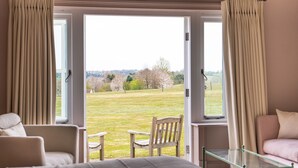 Een kamer met een bed, twee stoelen, een glazen tafel en uitzicht op een grasveld door open ramen.