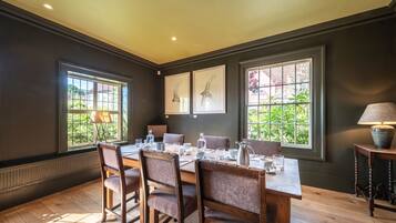 A dining room with a wooden table set for six, flanked by large windows with views of greenery.