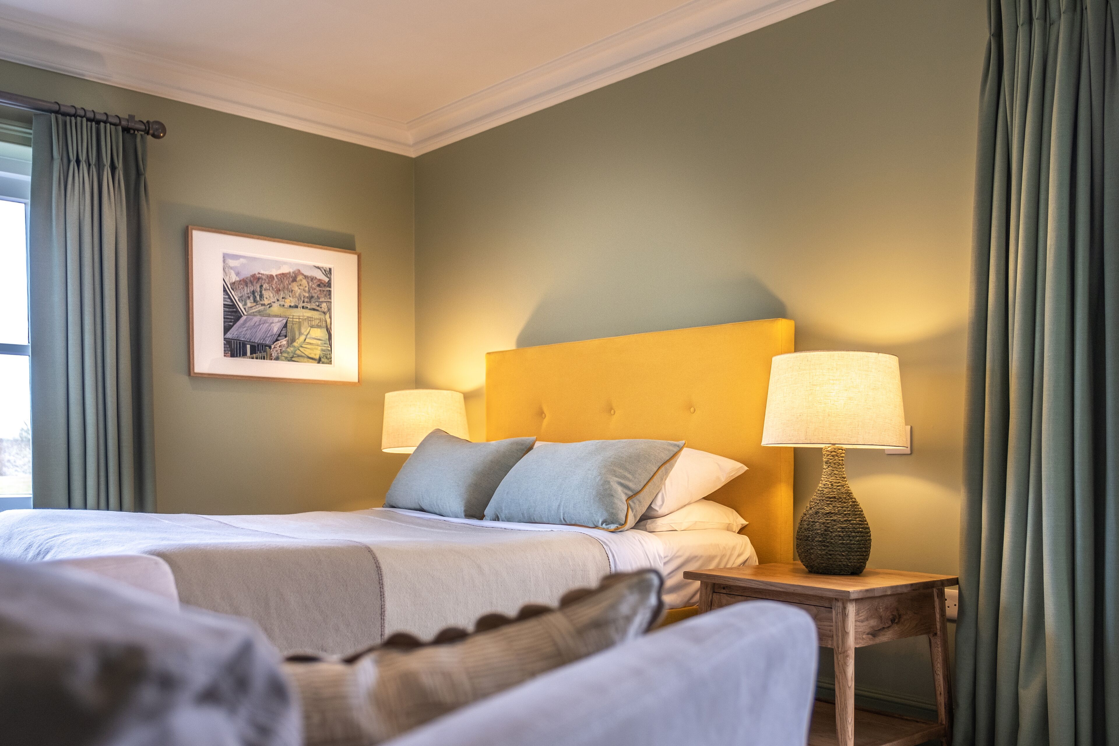 Une chambre d’hôtel avec un grand lit, deux lampes de chevet, un tableau encadré au mur et une fenêtre avec des rideaux.