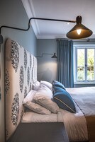 Un dormitorio con cabecera estampada, cama con ropa de cama blanca y azul, lámpara colgante y ventana con cortinas.
