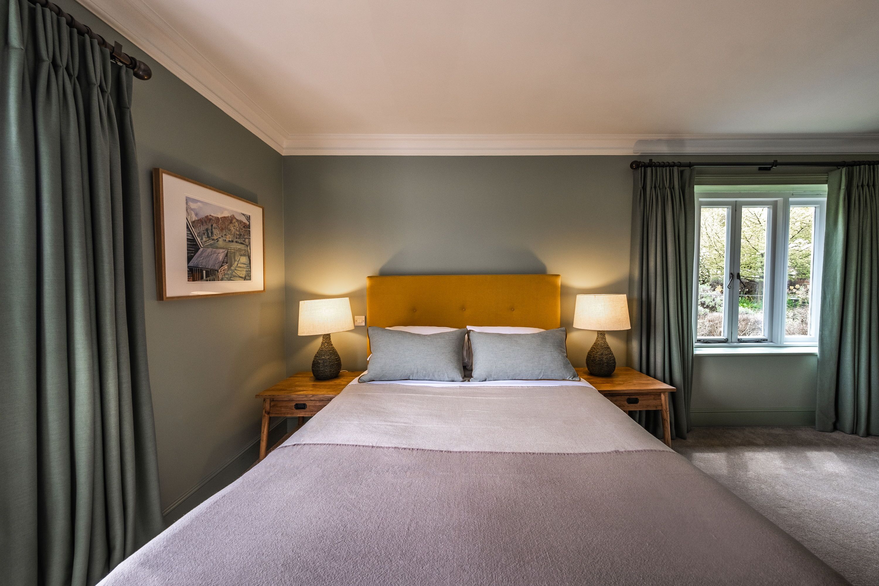A neatly made bed with a headboard, two bedside lamps, a framed picture on the wall, and a window with curtains.