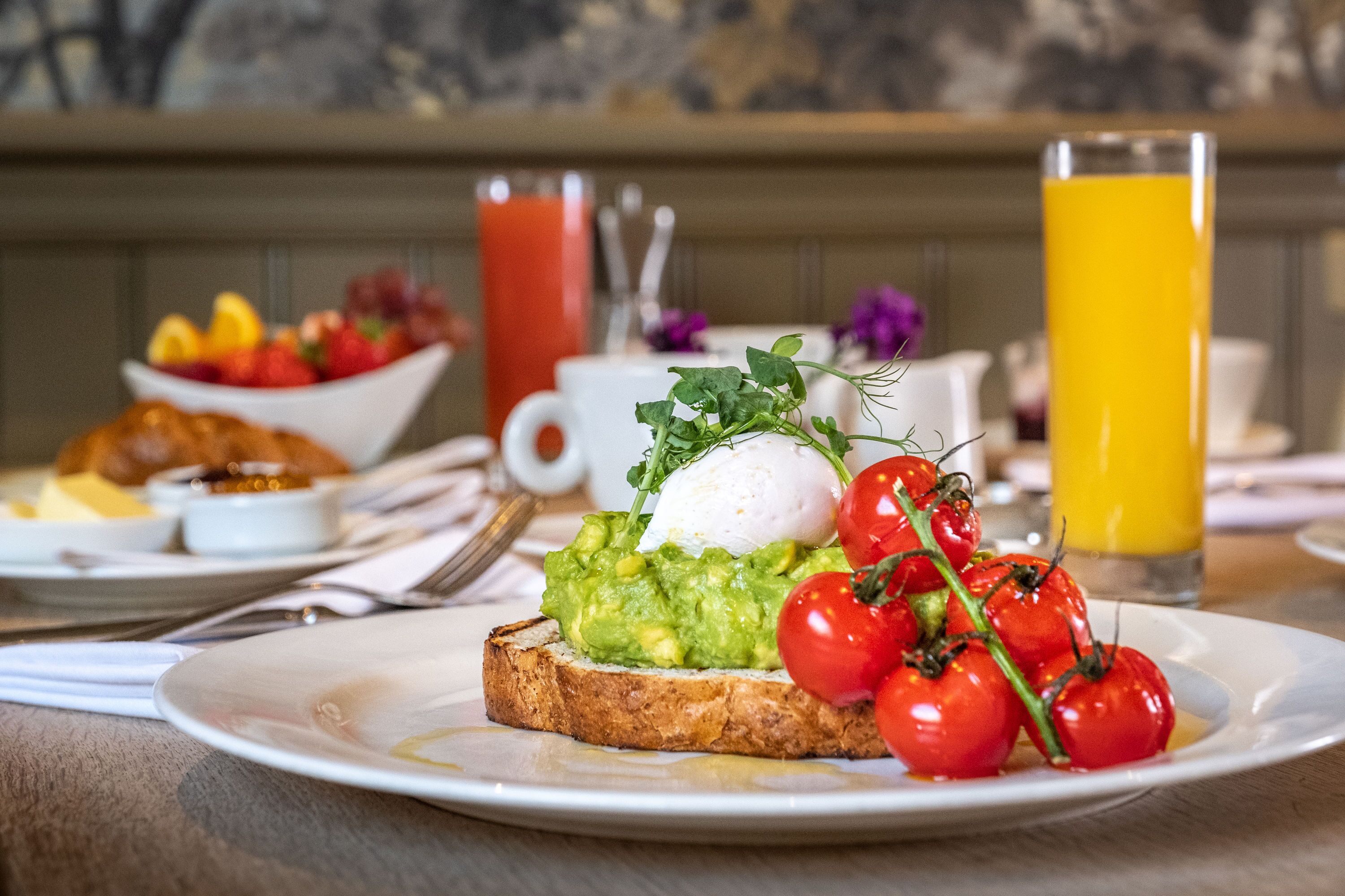 Un desayuno con tostadas cubiertas de aguacate, un huevo poché y tomates cherry. También hay tazones de fruta y un vaso de jugo de naranja.