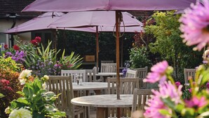 Een terras met houten tafels en stoelen, omringd door weelderige bloembedden en een paarse parasol.