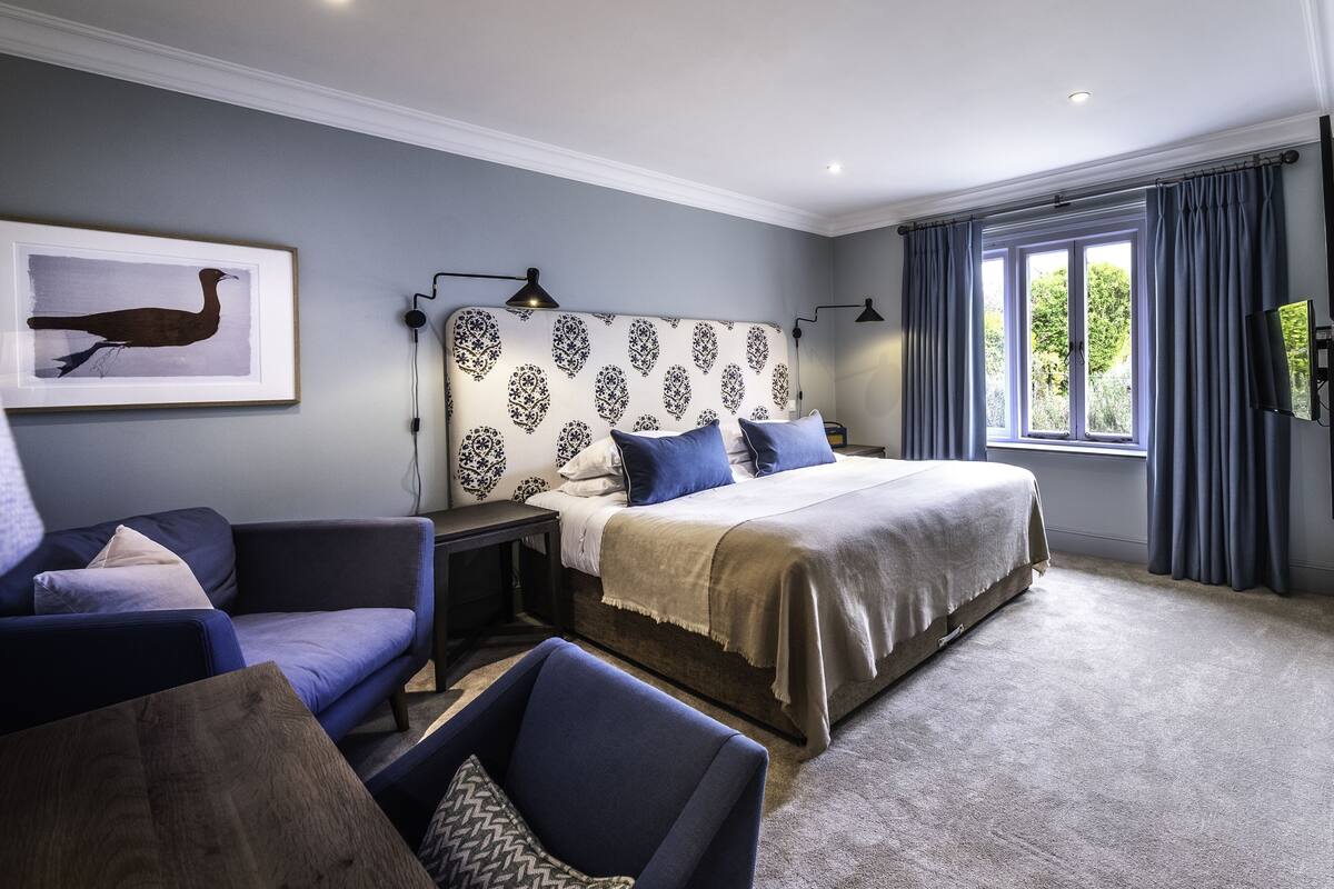 Een slaapkamer met een bed, een bank, een stoel, een nachtkastje, een lamp, een lijst met een afbeelding aan de muur en een raam met gordijnen.