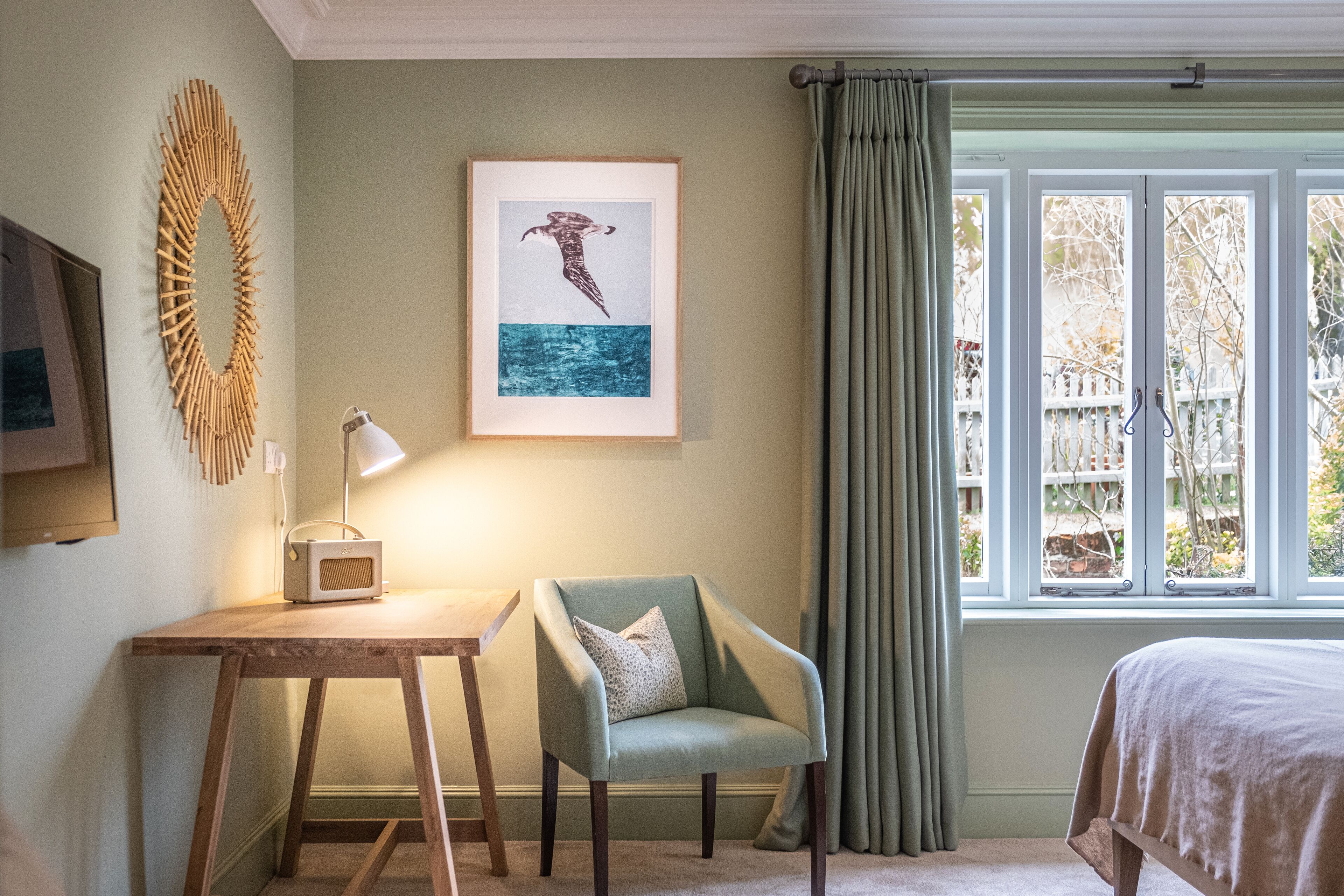 A bedroom with a bed, a chair, a desk with a lamp, a framed picture on the wall, and a window with curtains.