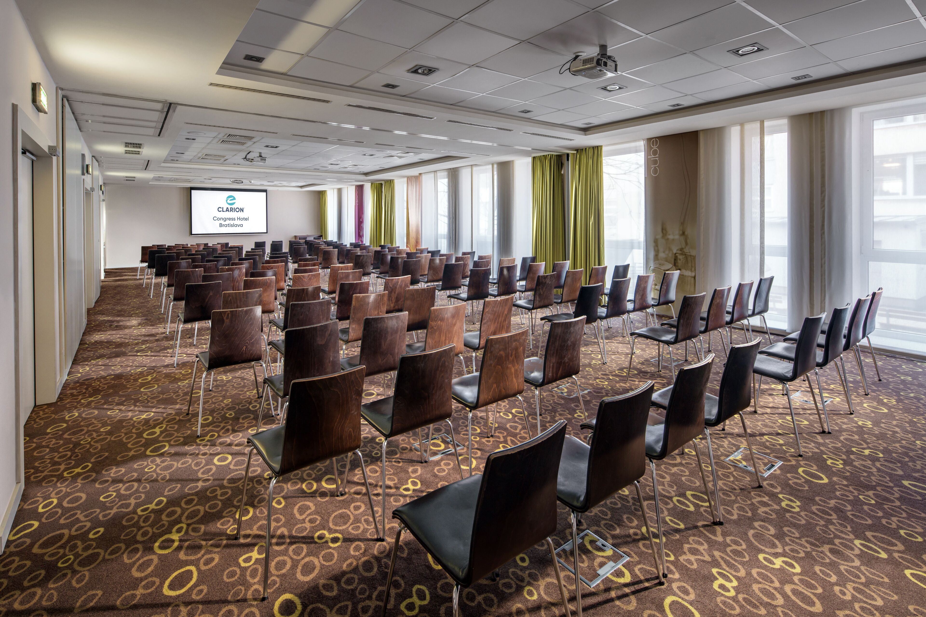 Une salle de conférence avec des rangées de chaises, un écran de projection et de grandes fenêtres.