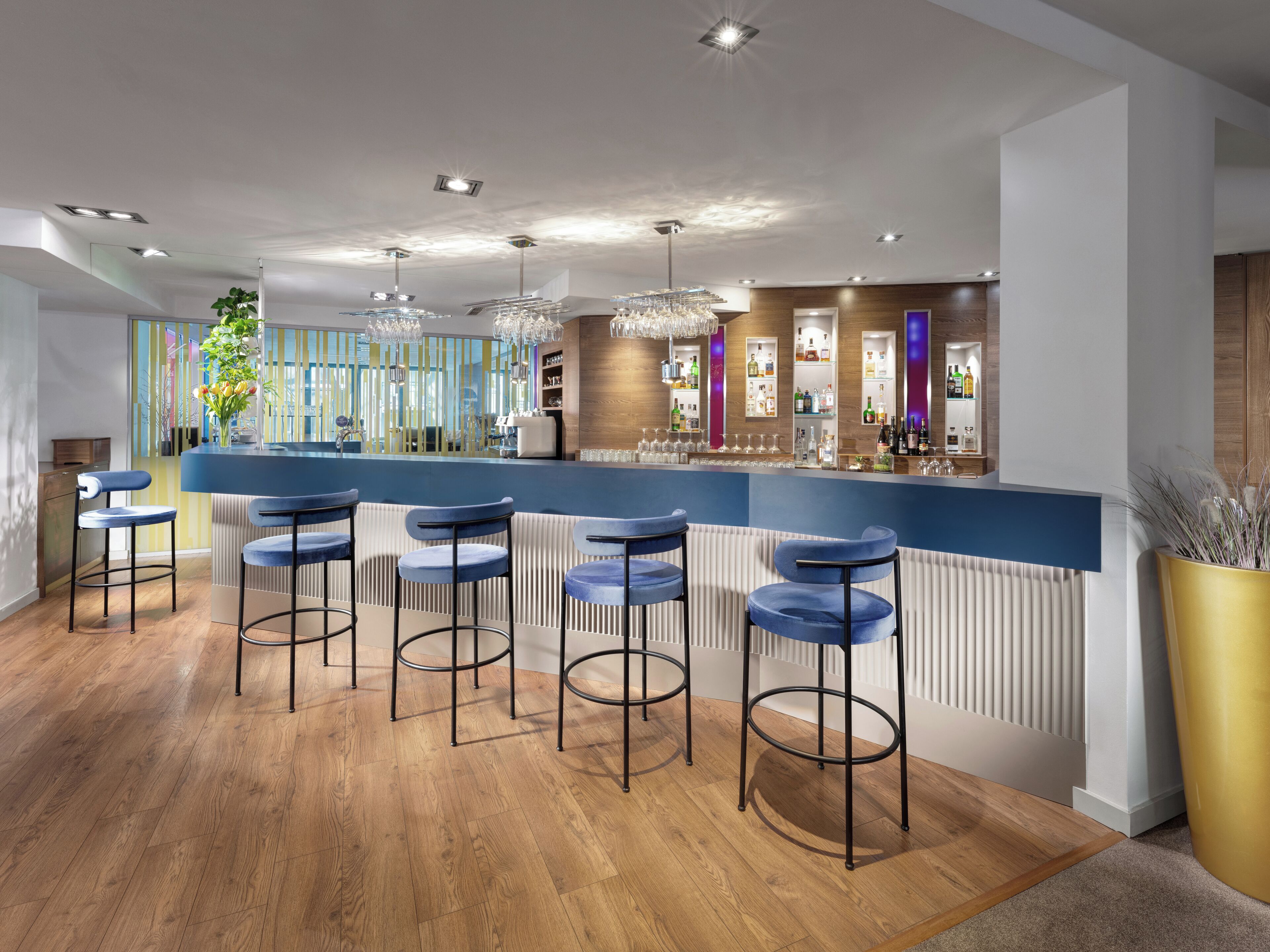 Eine moderne Bar mit Holzboden, blauen Barhocker und einer Auswahl an Flaschen und Gläsern auf Regalen.
