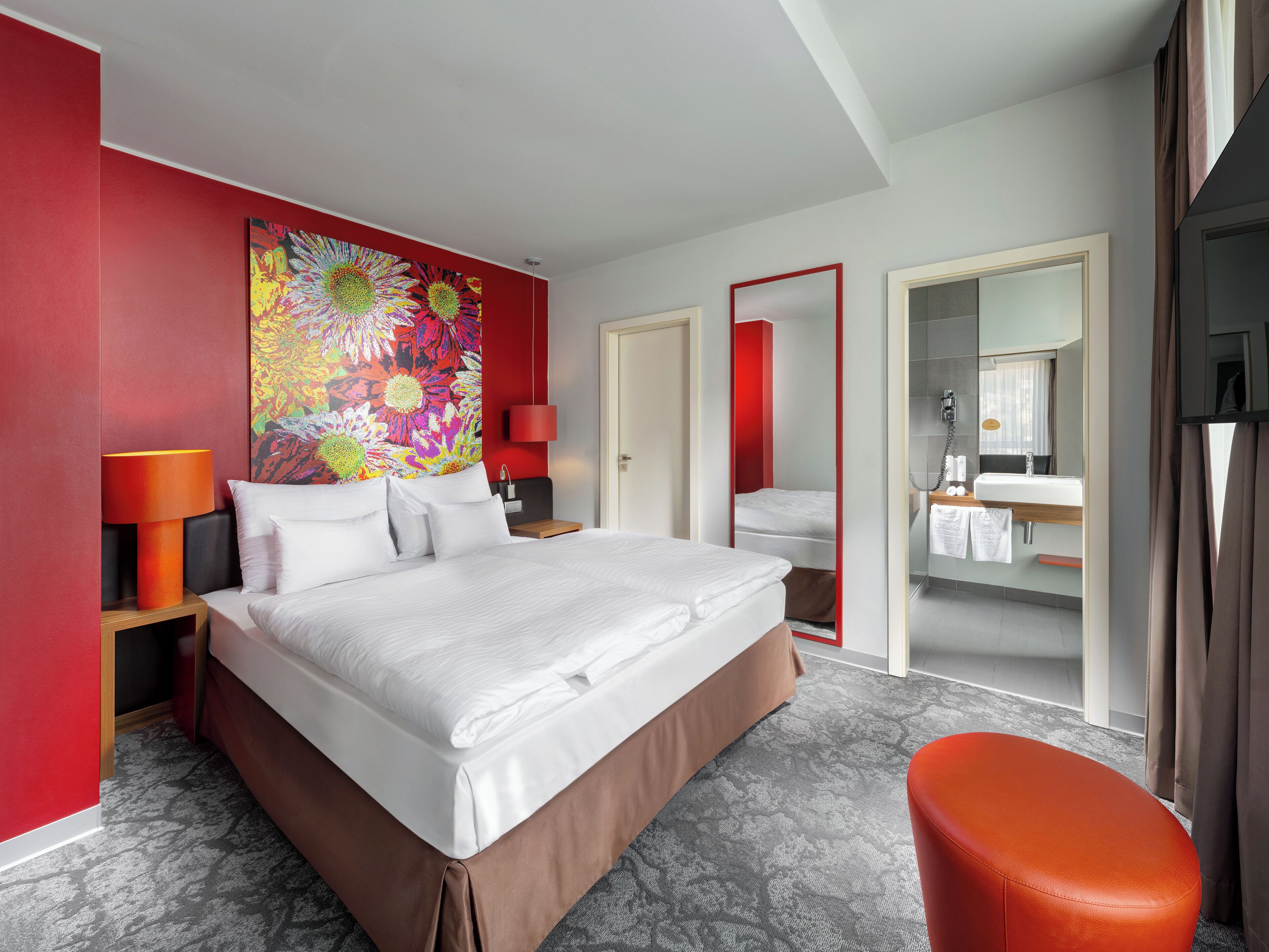 A hotel room with a large bed, a red accent wall, a colorful abstract painting, a red lamp, a bathroom with a mirror, and a red ottoman.