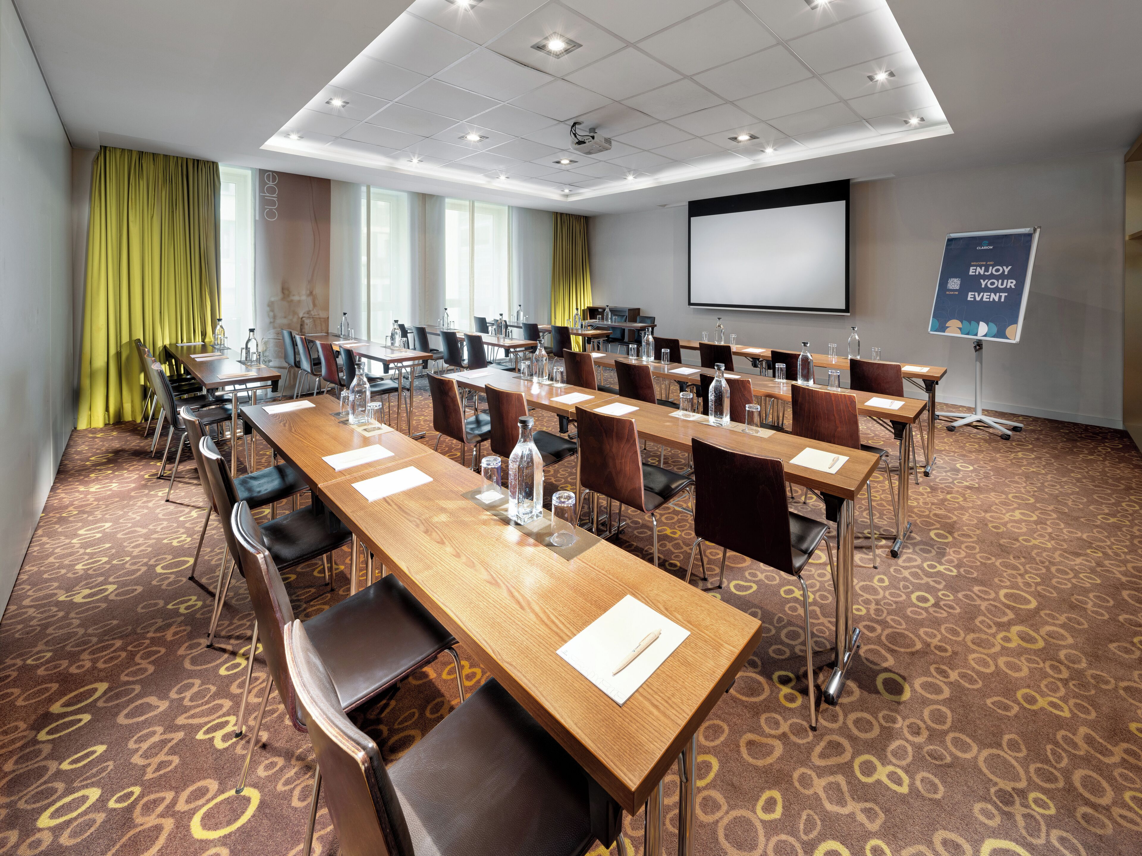 Un salón de conferencias con varias mesas y sillas, una pantalla de proyector y una pizarra para presentaciones.