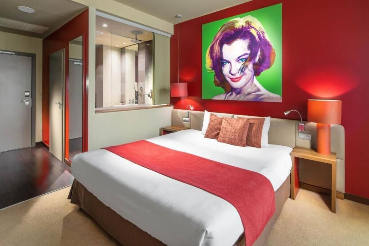 A hotel room with a large bed, a red accent wall, a painting, and a nightstand with a lamp.