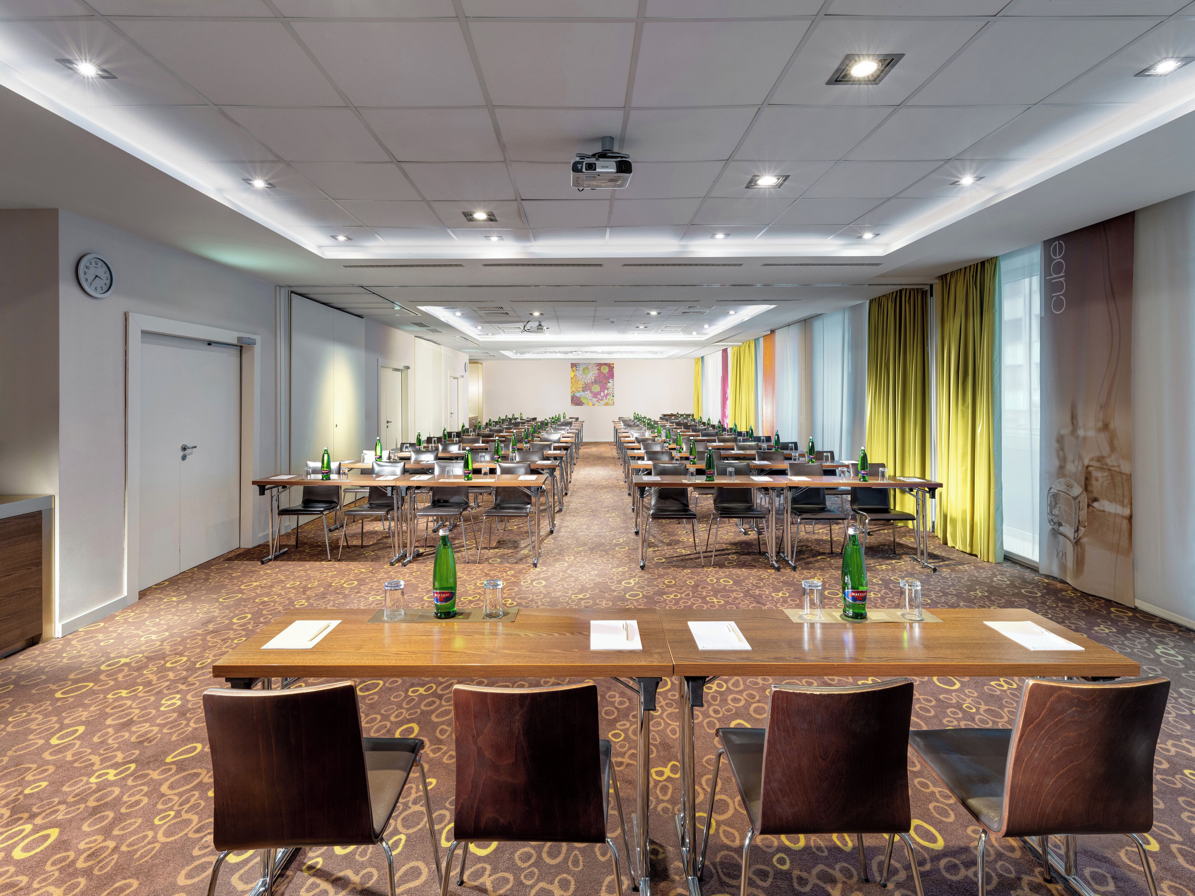 A conference room with multiple tables and chairs, equipped with water bottles and glasses, and a clock on the wall.