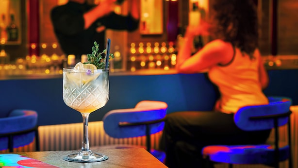 Un bar con un cóctel sobre una mesa, una persona al fondo y una barra con botellas.