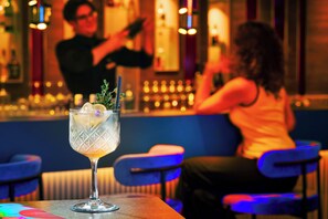 Een barsetting met een cocktail op een tafel, een persoon op de achtergrond en een bar met flessen.