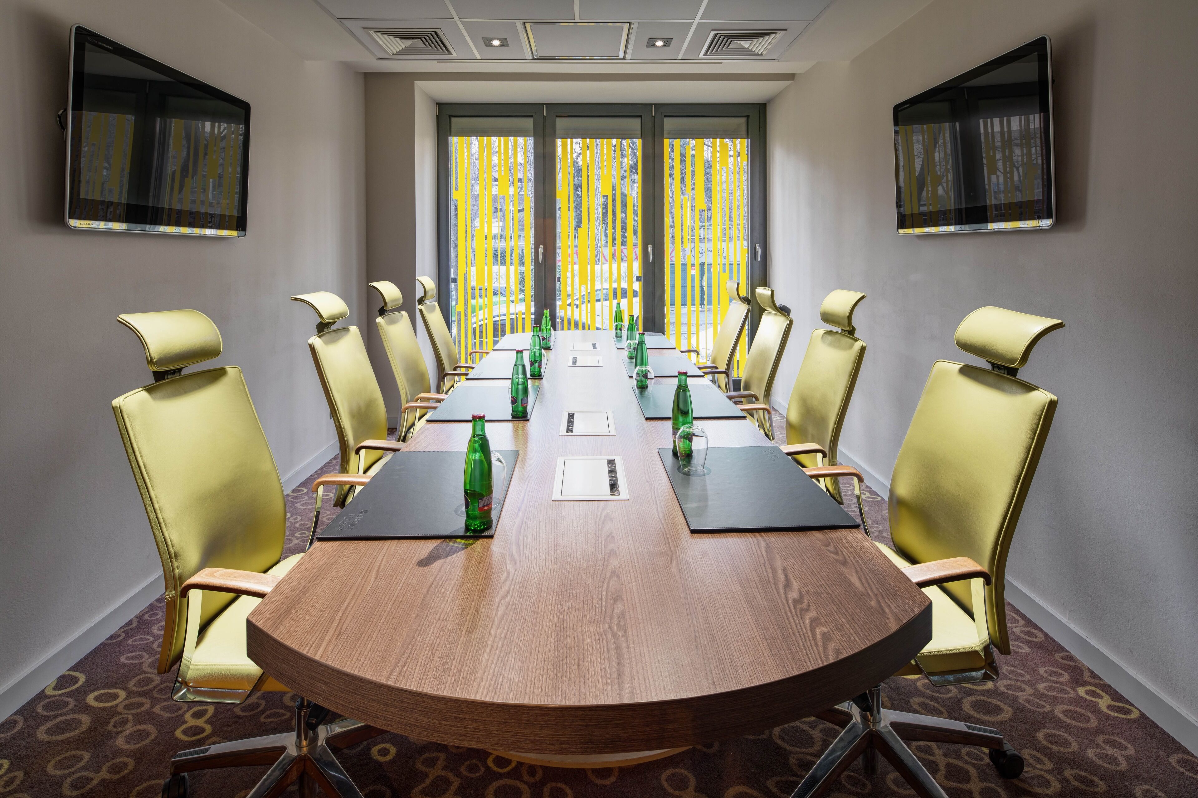 Uma sala de conferências com uma mesa comprida, cadeiras amarelas e grandes janelas com persianas verticais.