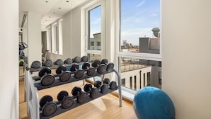 Un gimnasio con un estante de mancuernas, una pelota de ejercicios azul y grandes ventanales con vistas a la ciudad.