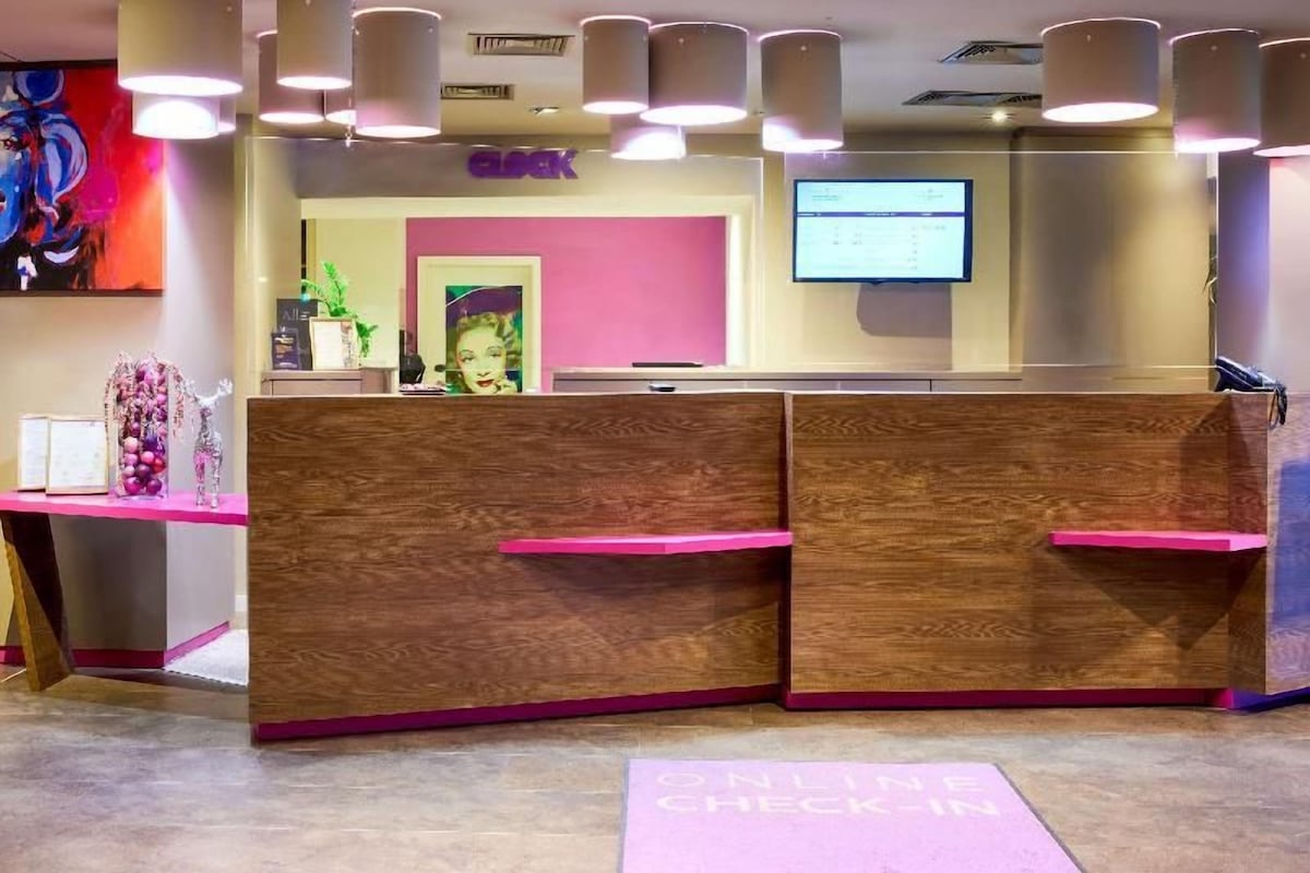 Un comptoir d’accueil au design rose et en bois, doté d’un écran numérique et agrémenté d’une plante.