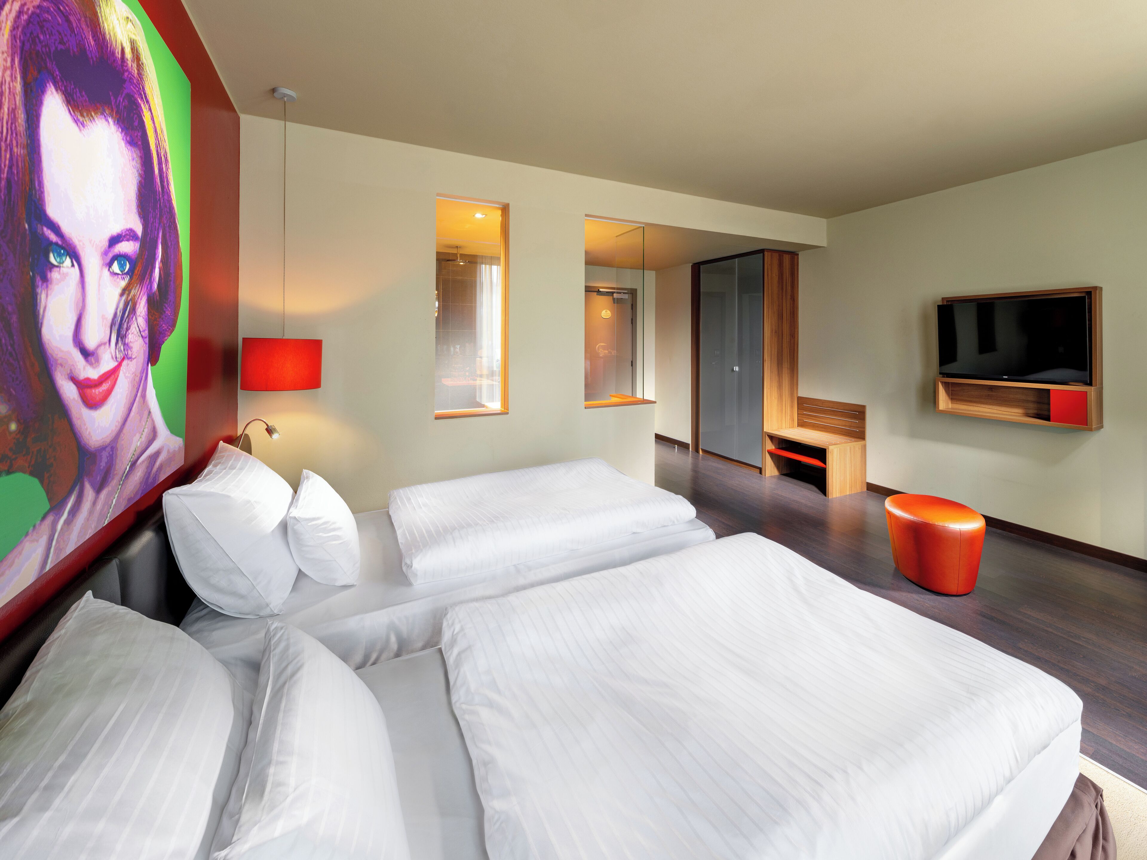 A hotel room with a large bed, a TV, a red stool, and a painting on the wall.
