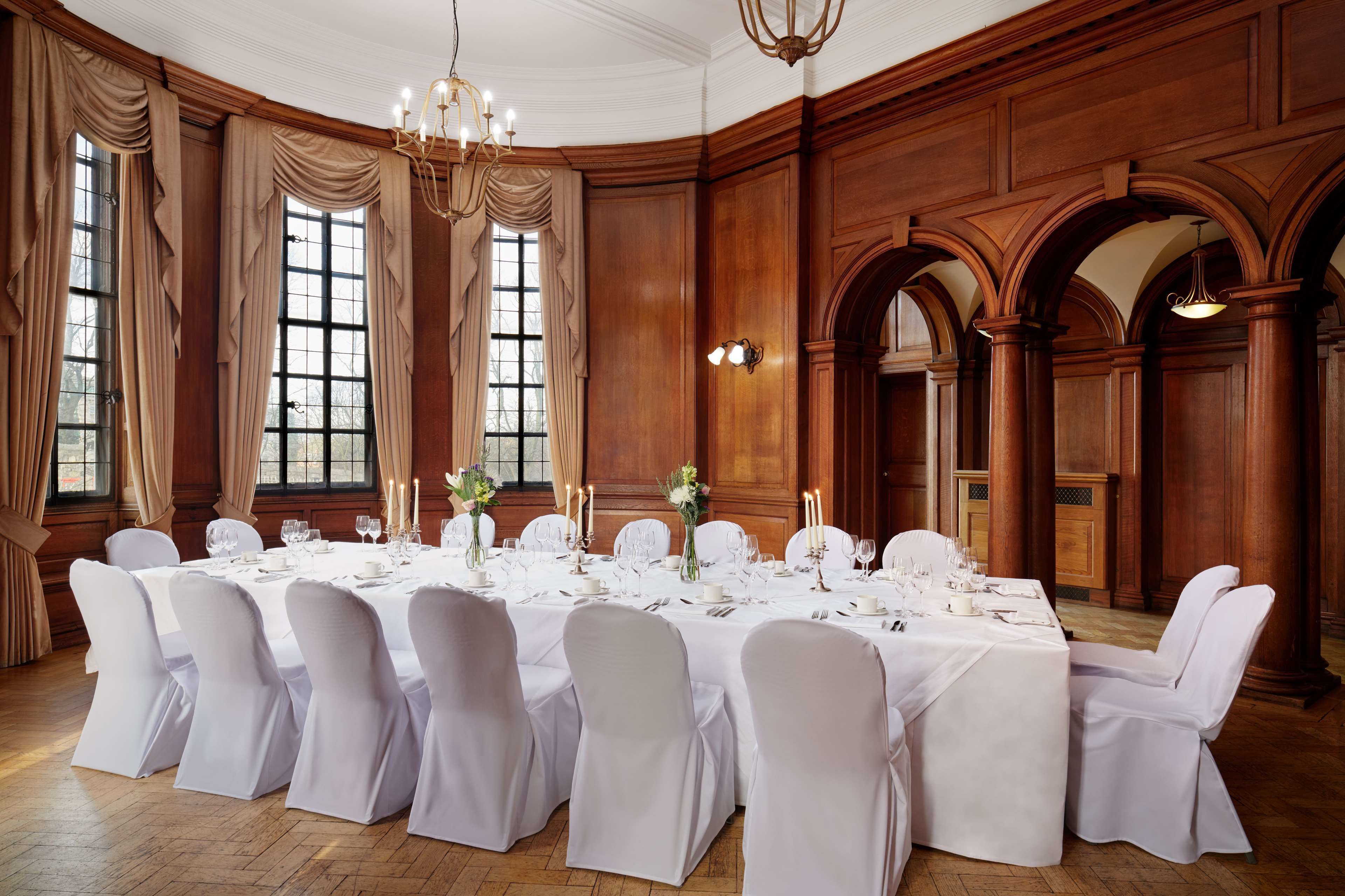A formal dining room with a long table set for a banquet, surrounded by wooden paneled walls and large windows.