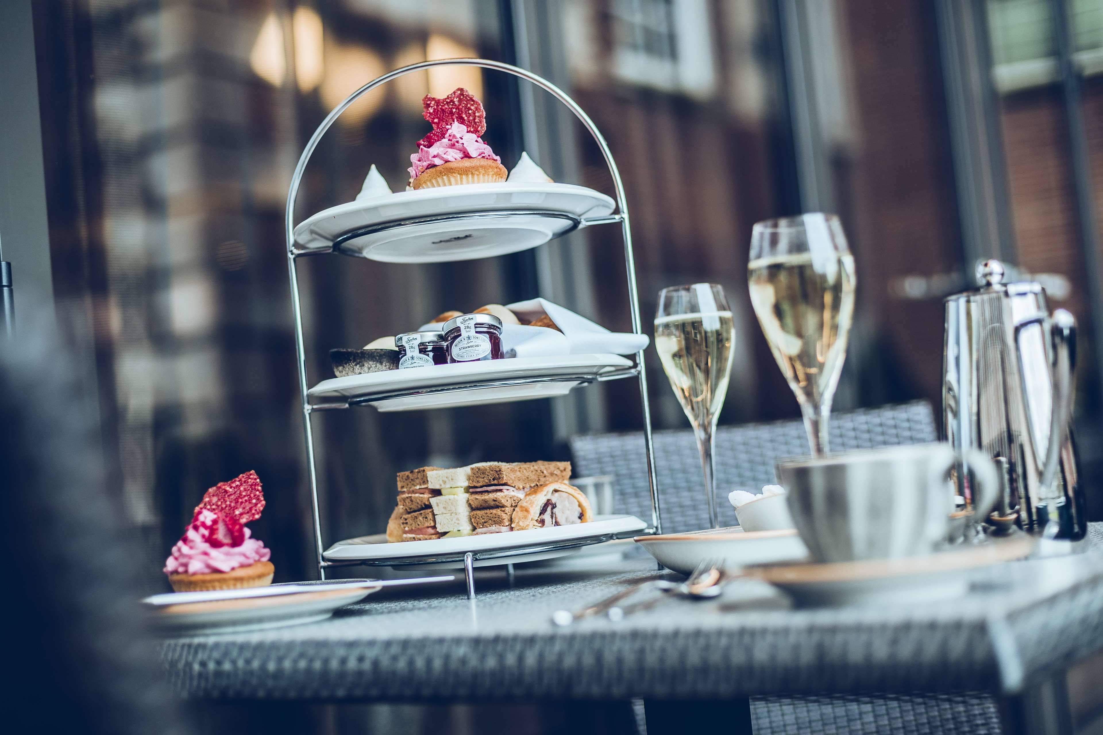 A three-tiered dessert stand with pastries and sandwiches, a glass of champagne, and a coffee pot on a table.