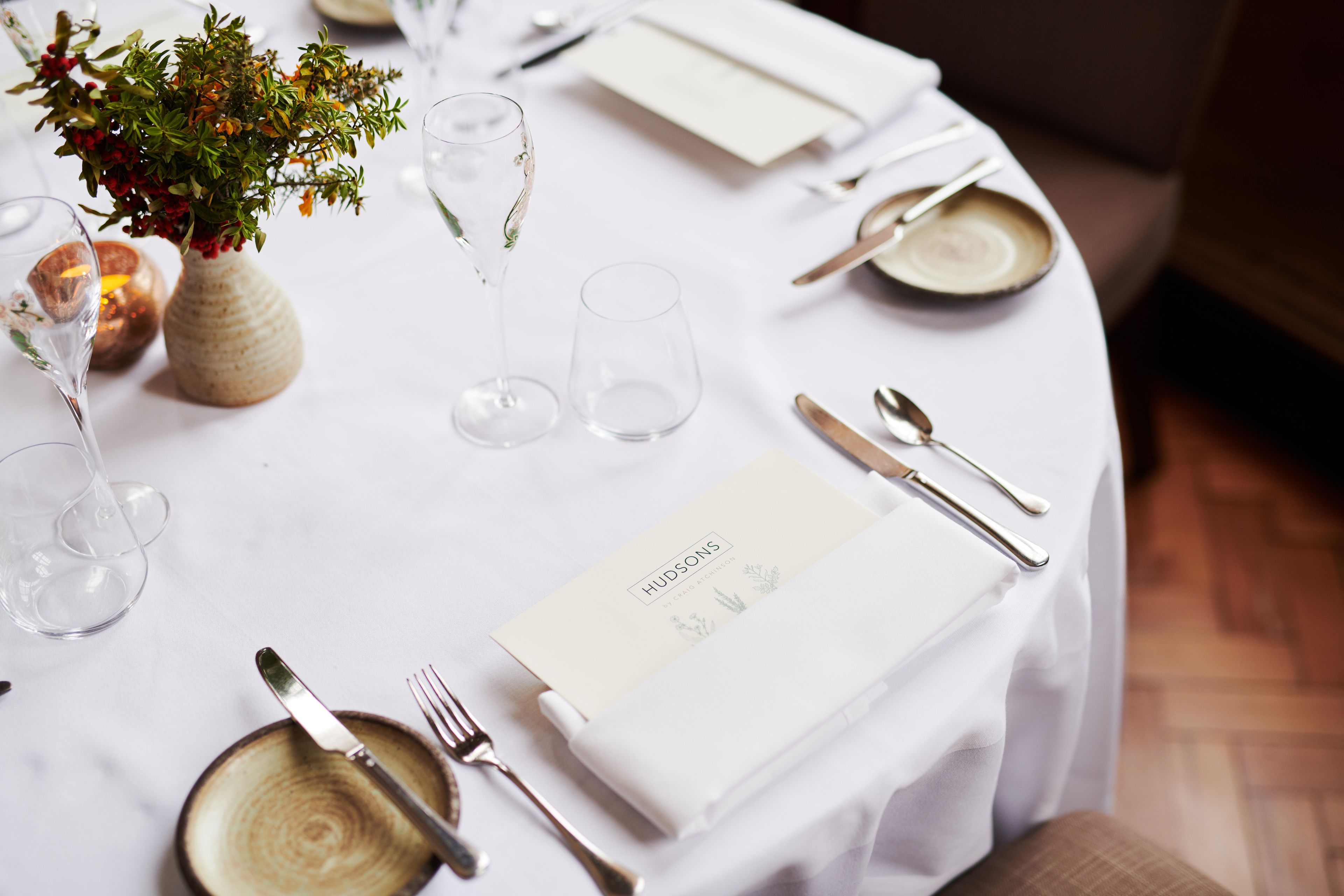 A neatly set dining table with a vase of flowers, wine glasses, and a menu card.
