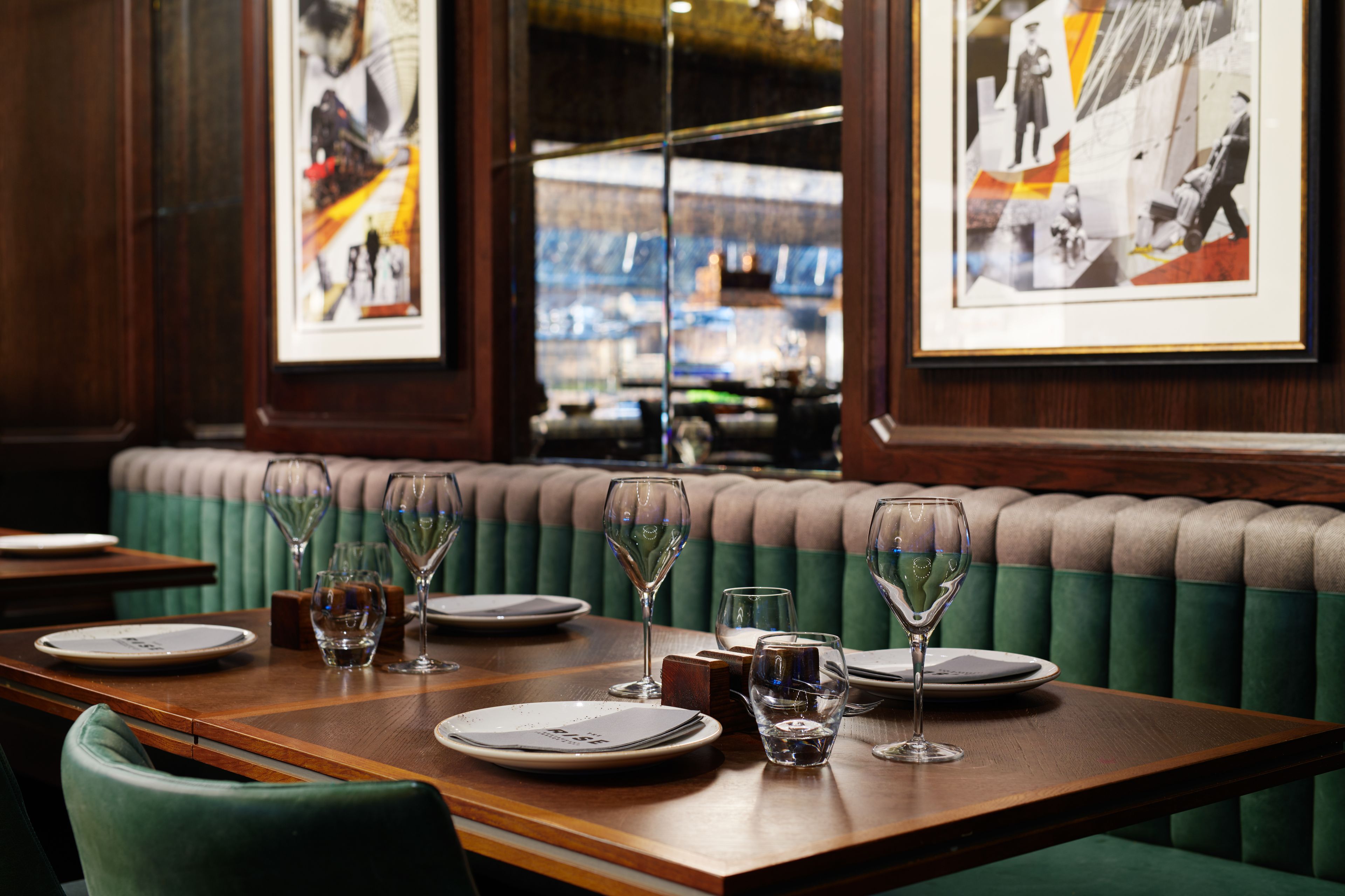 Interior de restaurante con una mesa de madera dispuesta para comer, un banco tapizado de color verde y cuadros en la pared.