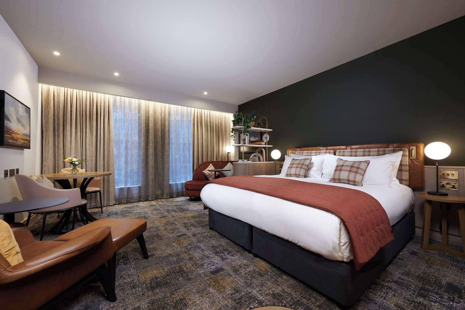 A modern hotel room with a large bed, a desk, a chair, and a bookshelf.