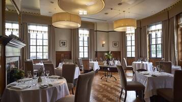 A formal dining room with multiple tables set for service, large windows with curtains, and a fireplace.