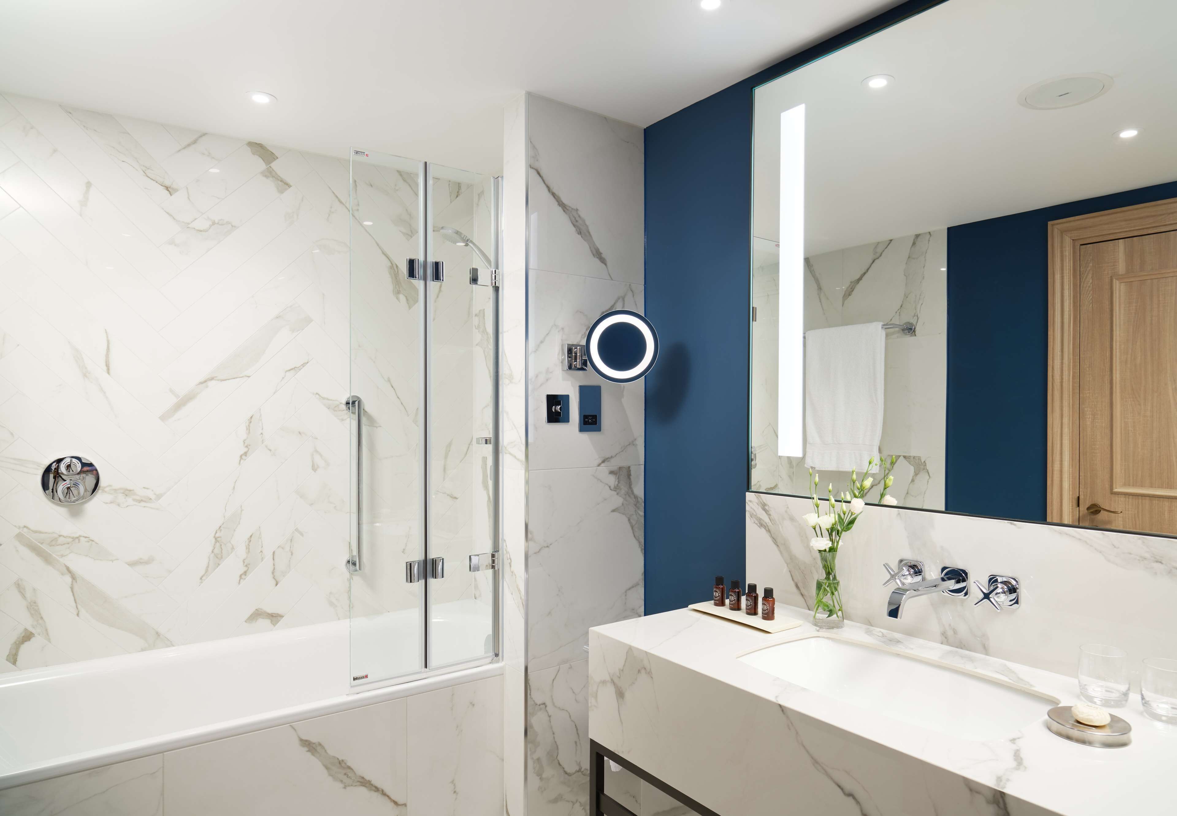 A modern bathroom with a glass-enclosed shower, a marble-tiled wall, a blue accent wall, a large mirror, and a vanity with a sink and toiletries.
