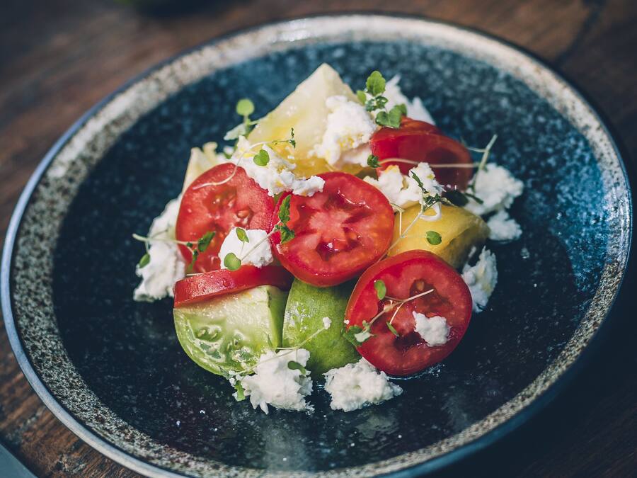 Une assiette de nourriture comprenant des tomates en tranches, du fromage et des légumes verts, le tout présenté sur une assiette foncée.