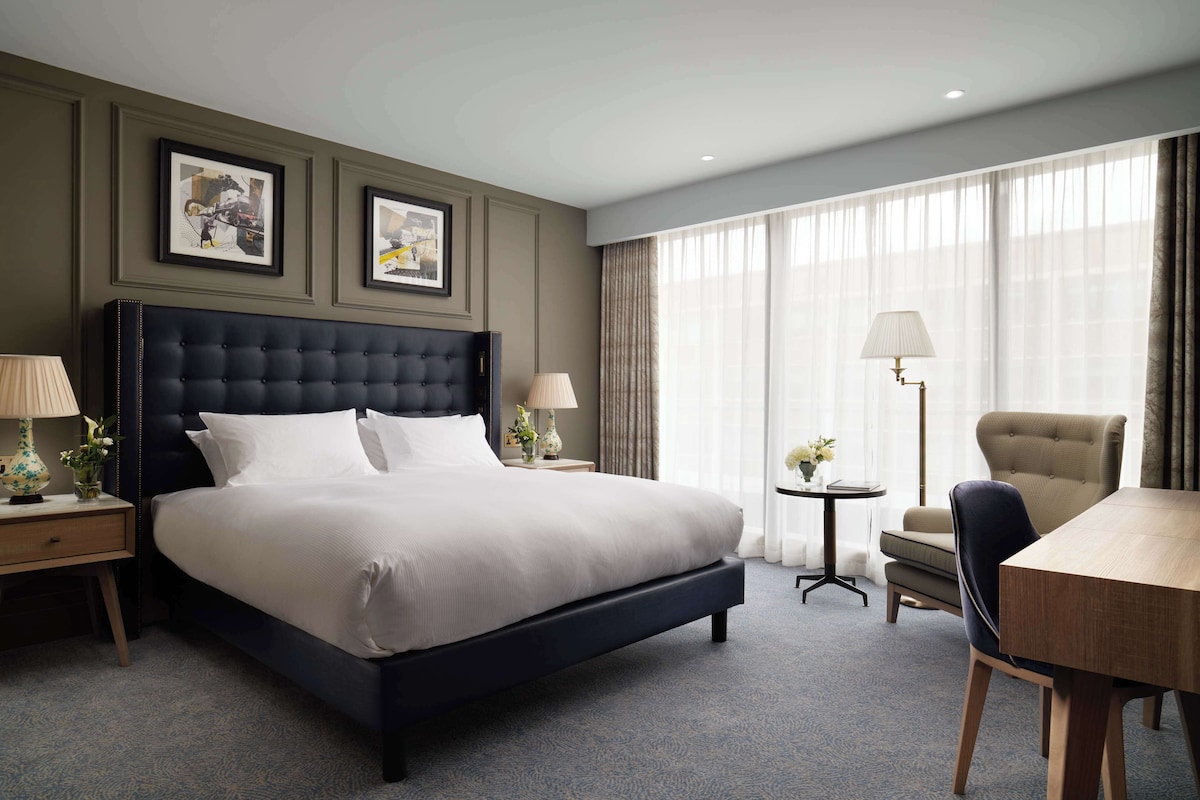 Habitación de hotel con una cama grande, un escritorio, una silla y una mesa. Hay dos cuadros en la pared, una lámpara en el escritorio y otra en la mesa.
