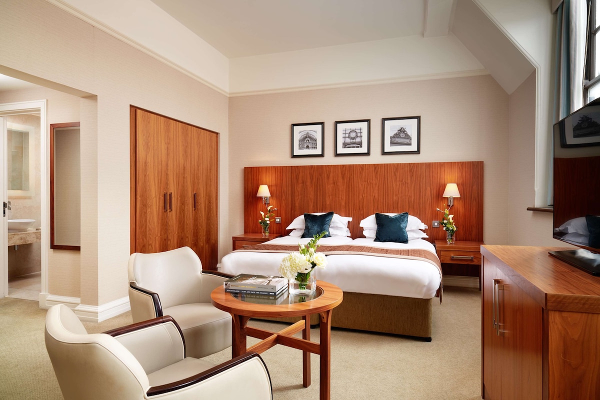A hotel room with a large bed, two bedside tables with lamps, a wooden wardrobe, a round wooden table with a vase of flowers, and a television on a wooden cabinet.