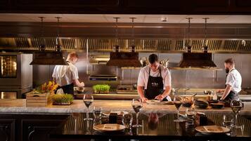 A professional kitchen with chefs preparing food, a marble countertop, and wine glasses.