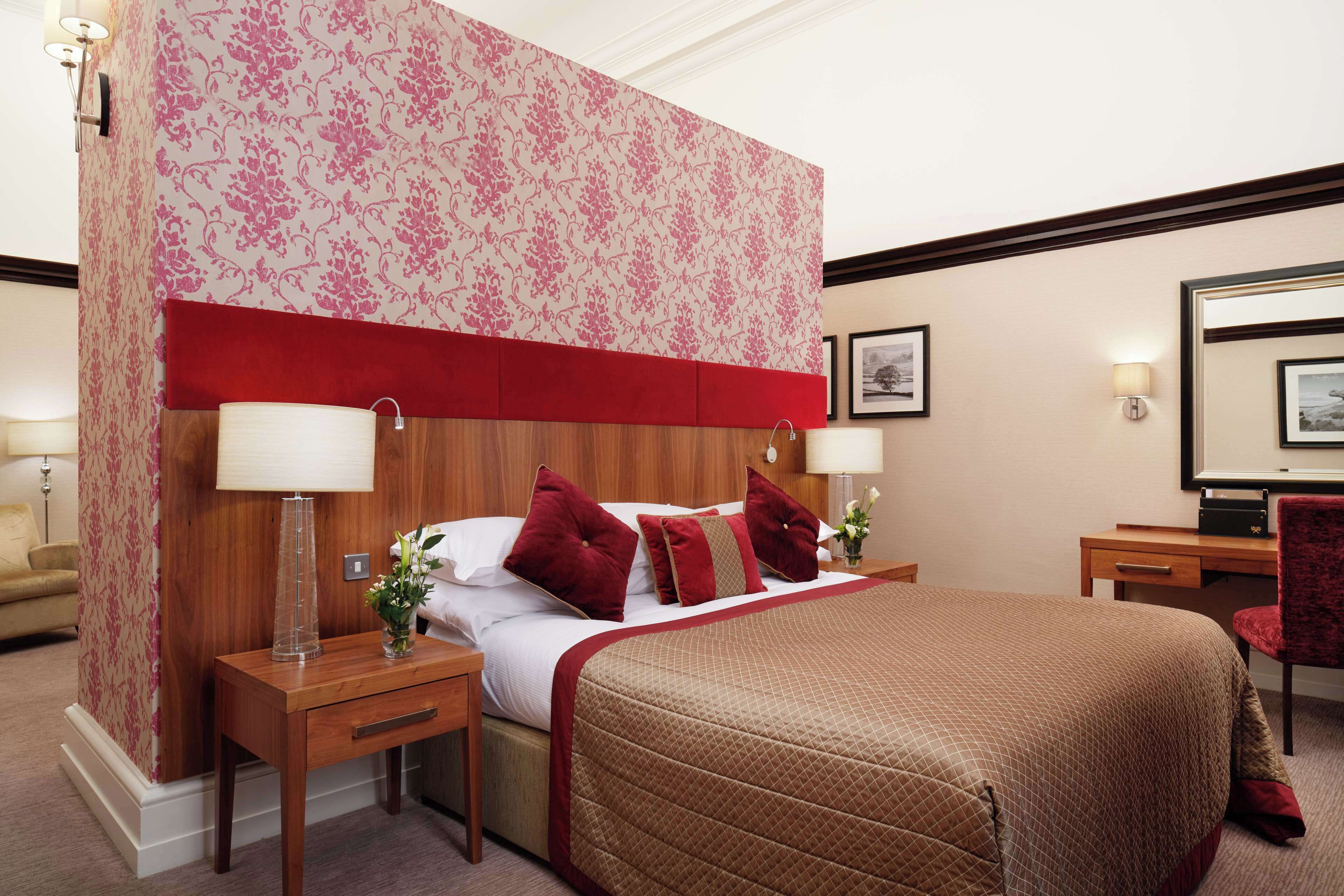 A hotel room with a large bed, two bedside tables, a red headboard, floral wallpaper, and a desk with a chair.