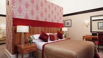 A hotel room with a large bed, two bedside tables, a red headboard, floral wallpaper, and a desk with a chair.