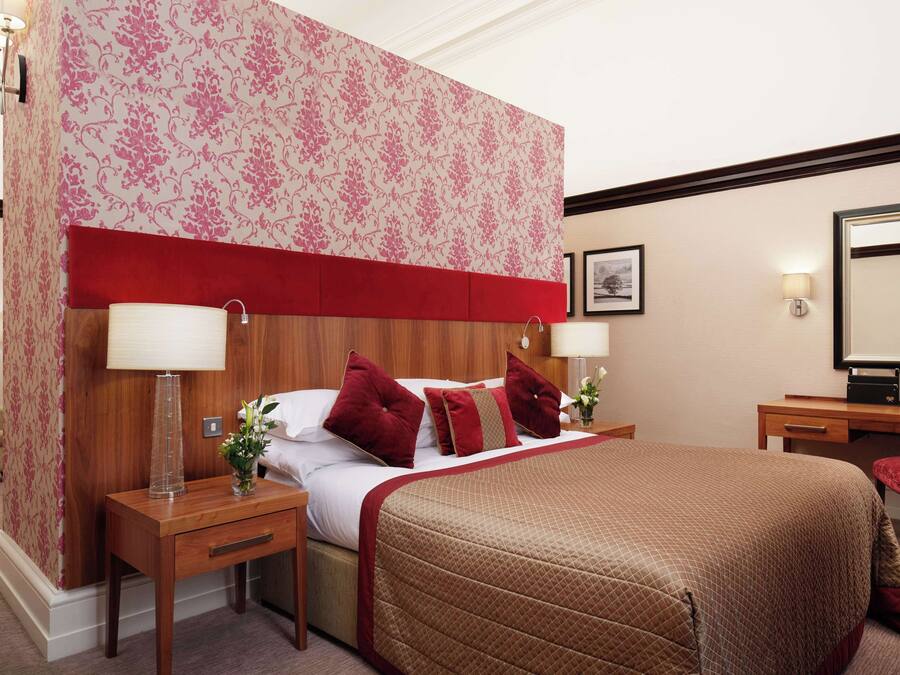 Habitación de hotel con una cama grande, dos mesitas de noche, un cabecero rojo, papel tapiz floral y un escritorio con una silla.