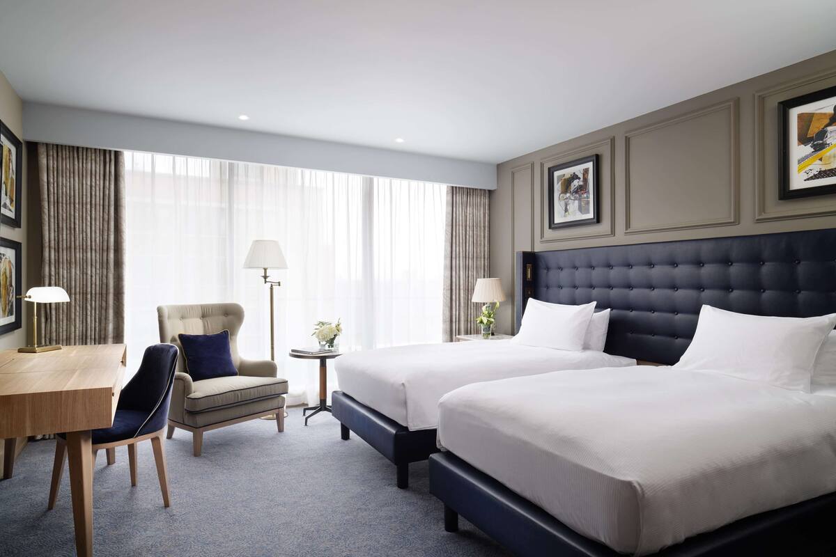 Hotellihuone, jossa on kaksi sänkyä, työpöytä, tuoli ja pieni pöytä, jossa on lamppu.
