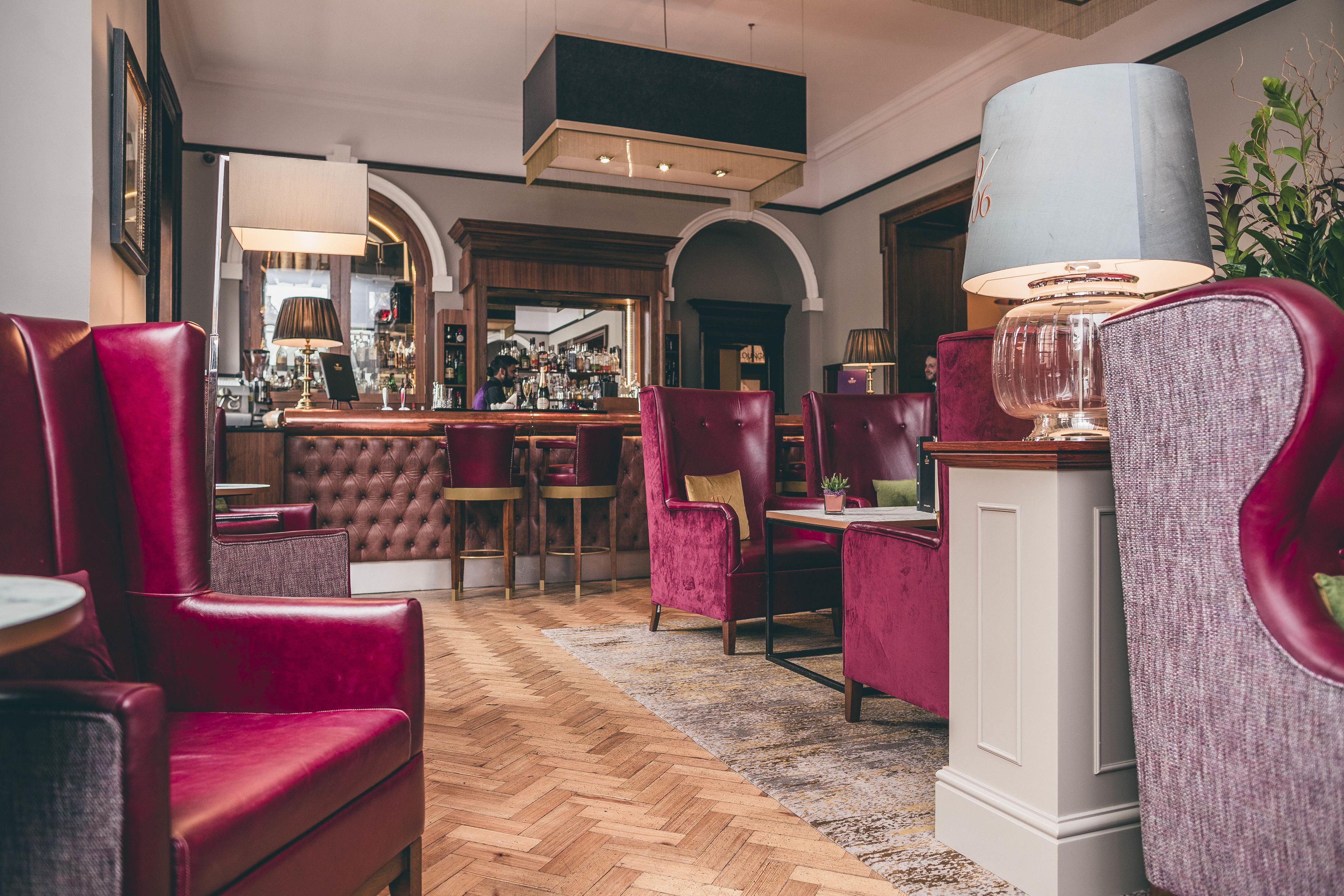 A bar area with red upholstered chairs, a wooden herringbone floor, and a bar counter with a bartender.