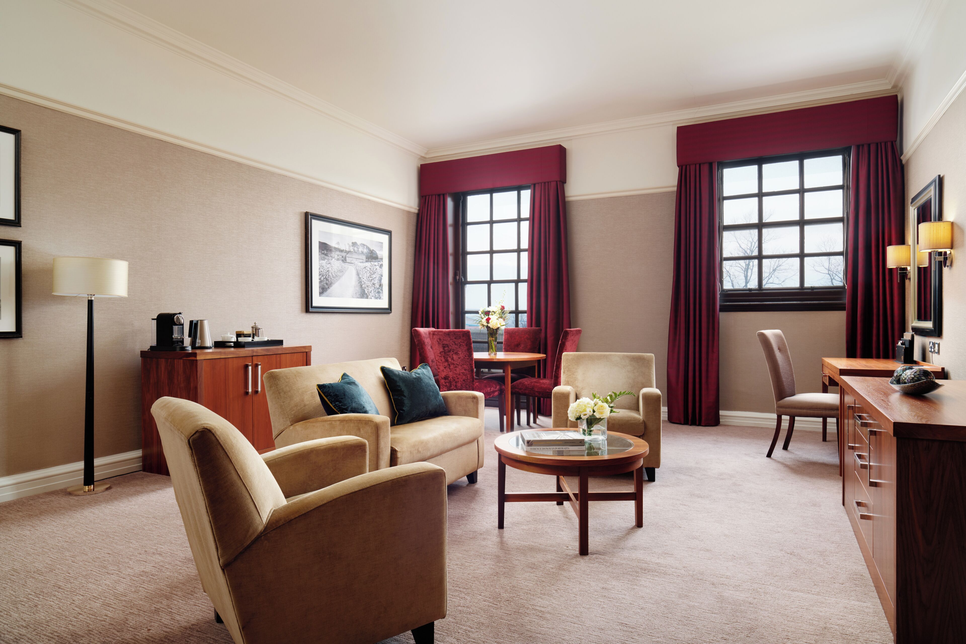 A hotel room with a sofa, armchairs, a coffee table, a dining table with chairs, a desk with a chair, a lamp, a framed picture, and a window with curtains.