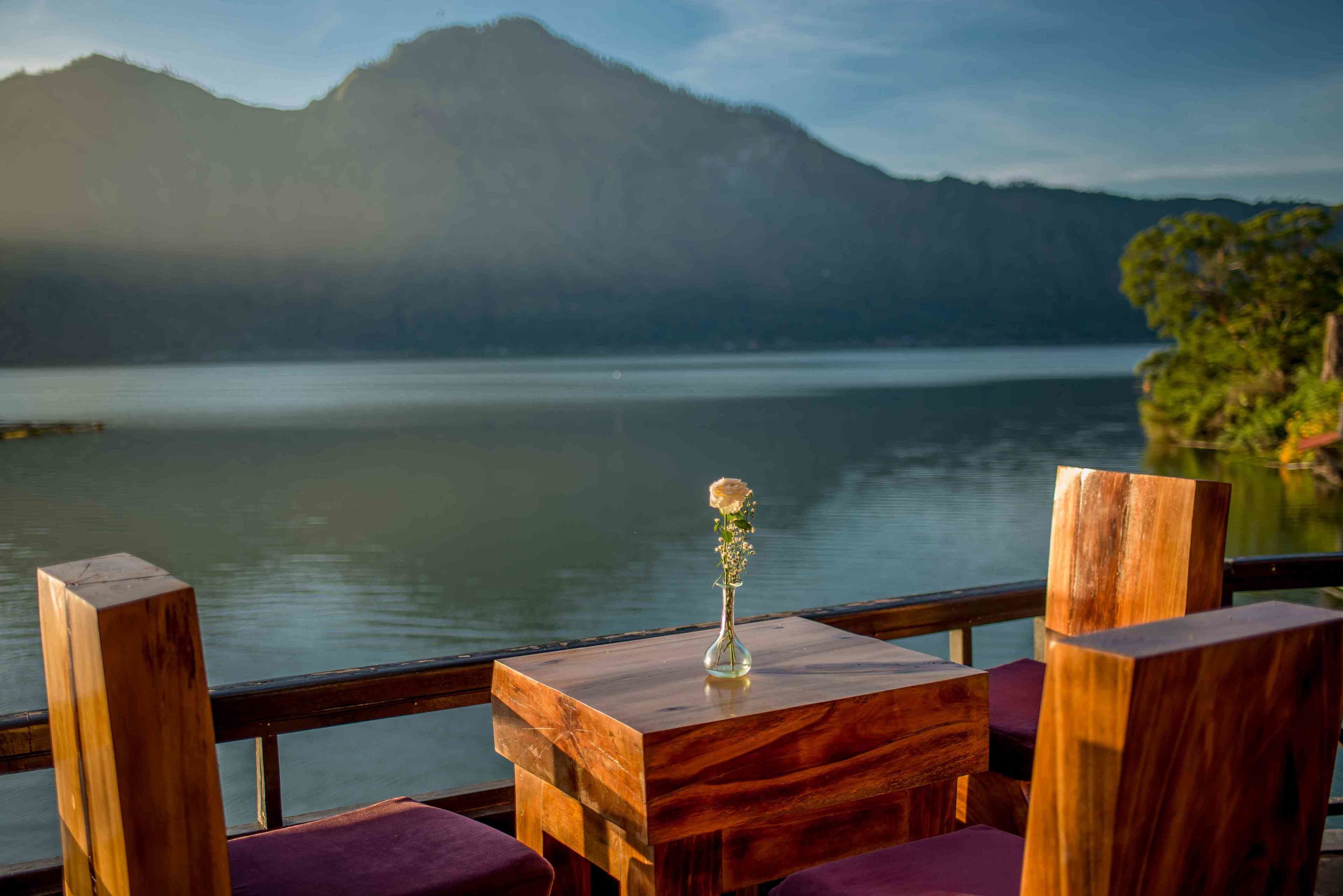 Una mesa de madera con un jarrón de flores, frente a un lago con montañas al fondo.