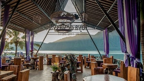 Un área de comedor al aire libre con mesas y sillas de madera, un colgante decorativo y vistas al mar y a las montañas.