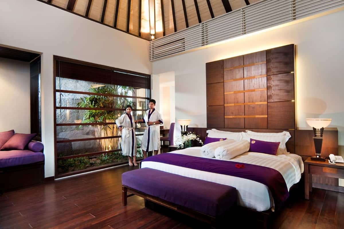Une chambre d’hôtel moderne avec un grand lit, une tête de lit en bois, une table de chevet avec une lampe et une vue sur la verdure par une grande fenêtre.
