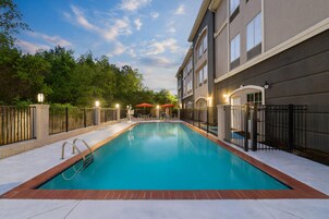 Una piscina rectangular con una escalera metálica, rodeada por un patio de ladrillo y una valla de metal.